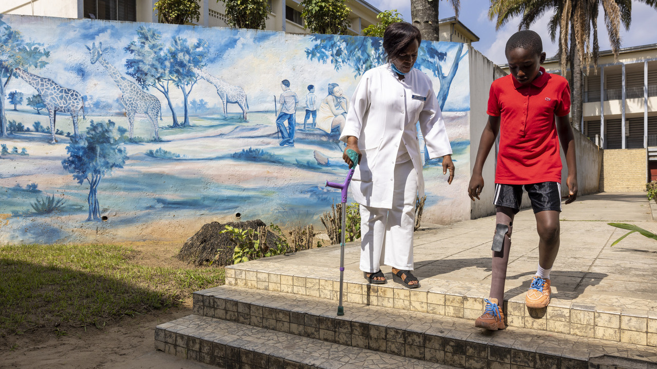 Ein kleiner Junge mit Beinprothese ist auf den Stufen vor dem Krankenhaus in Kinshasa zu sehen. Neben ihm steht seine Physiotherapeutin, die Krücken bereithält.